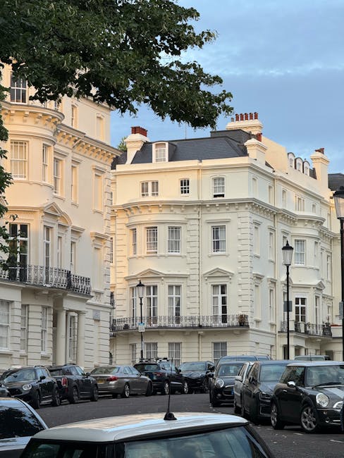 A row of elegant white Victorian-style residential buildings with ornate architectural details and bay windows, situated on a street with parked cars along the pavement. In the foreground, a collectio