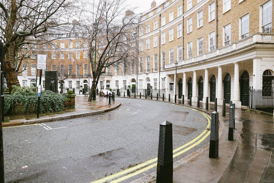 A row of elegant white Victorian-style residential buildings with ornate architectural details and bay windows, situated on a street with parked cars along the pavement. In the foreground, a collectio
